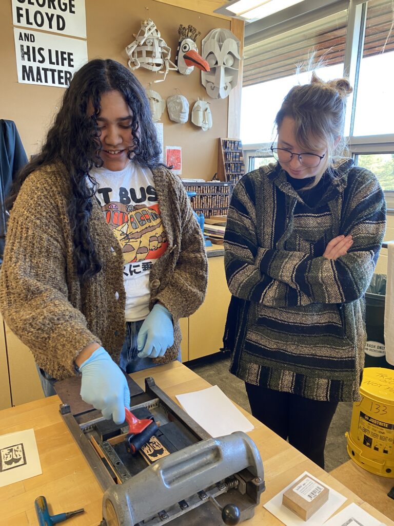 Two students in an art studio using a proof press, surrounded by art-in-progress, art supplies, masks, and a poster that says "George Floyd and His Life Matter."