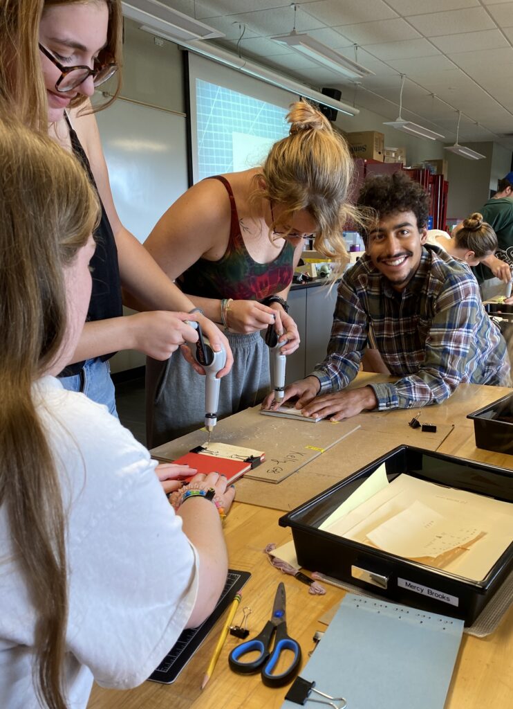 Students using manual drills on their books-in-progress, assisted by classmates, to prepare for binding.