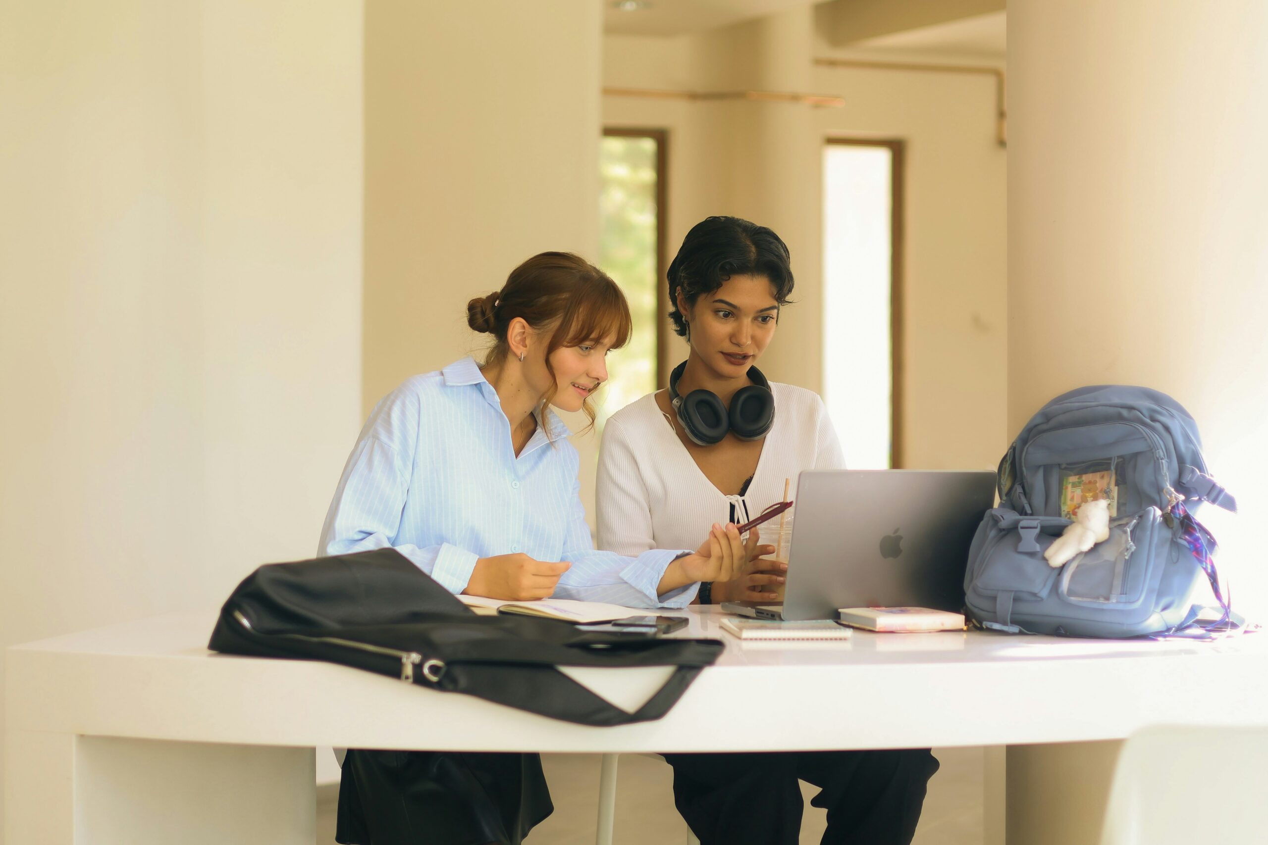 Two women sitting at a desk looking at a laptop. Books, a backpack, and a handbag are on the desk.