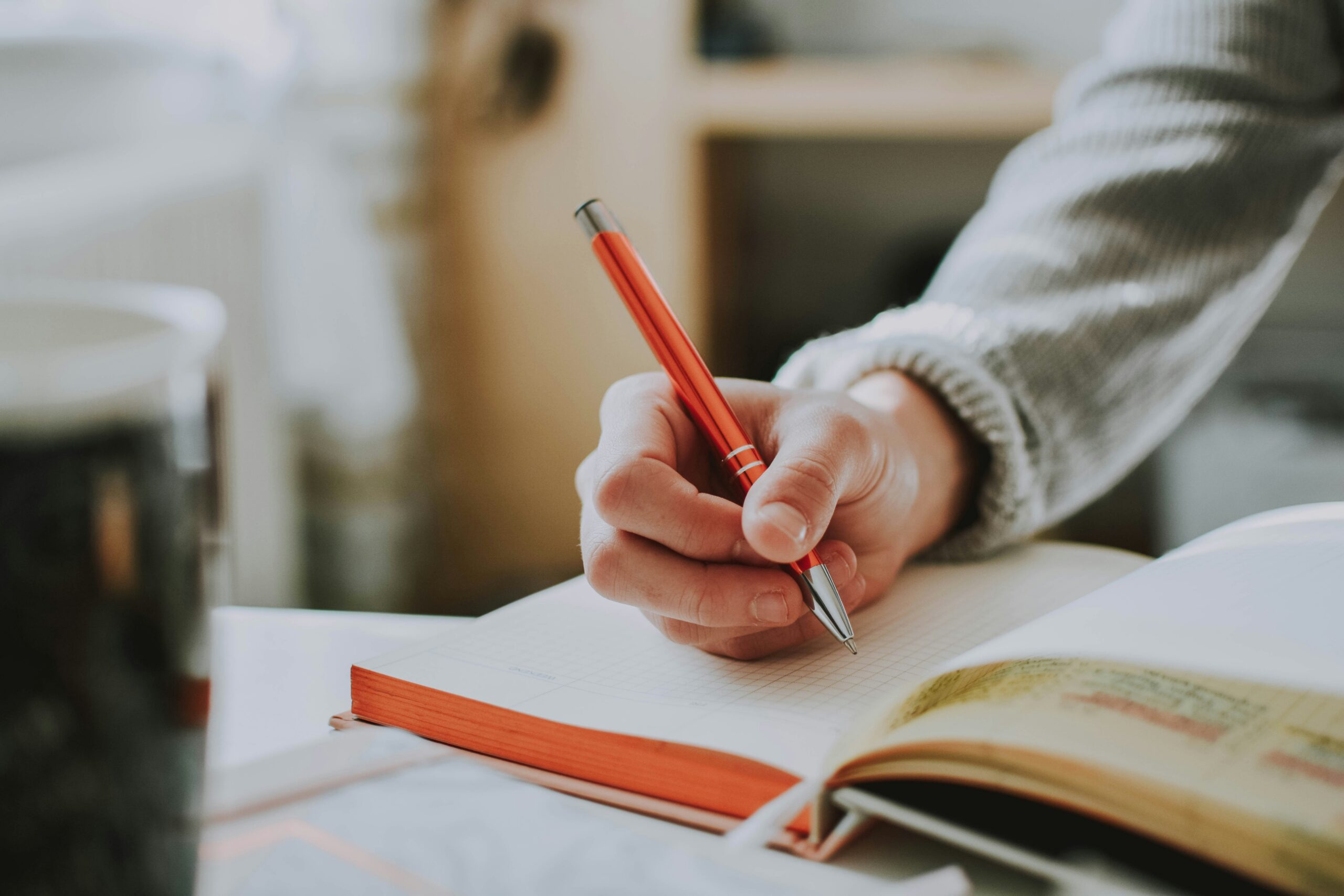 A person using an orange pen to write in an open note book.